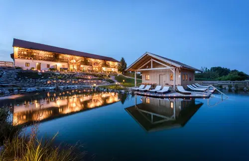 Ein modernes Baumhaus steht am Ufer eines ruhigen Teichs, w&auml;hrend die Fassade des angrenzenden Geb&auml;udes im Abendlicht erleuchtet ist. Die Wasseroberfl&auml;che spiegelt die Architektur und die umliegende Landschaft wider.