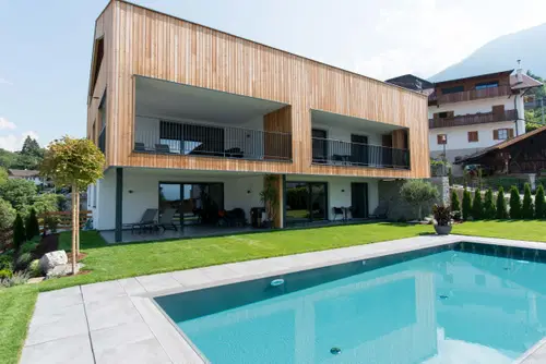 Modernes Haus mit Holzverkleidung und gro&szlig;en Fenstern. Vorne ein Schwimmbecken und eine gepflegte Rasenfl&auml;che. Im Hintergrund sind Berge und weitere Geb&auml;ude sichtbar.