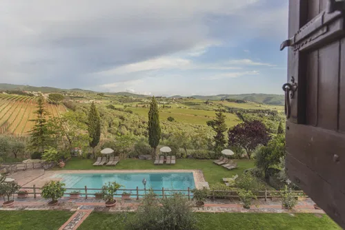 Aussicht auf einen Swimmingpool mit Liegen, umgeben von &uuml;ppigem Gr&uuml;n und Weinbergen in der Toskana. Der Himmel ist bew&ouml;lkt mit Sonnenstrahlen, die durchscheinen.
