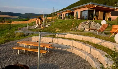 Moderne Holzh&auml;user auf einer Anh&ouml;he, umgeben von einer gr&uuml;nen Rasenfl&auml;che. Stufen aus Granit f&uuml;hren zu einer Grillstelle mit Holztischen und Liegest&uuml;hlen. Im Hintergrund sind sanfte H&uuml;gel und ein klarer Himmel zu sehen.