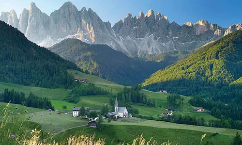 Eine malerische Landschaft mit einer kleinen Kirche und mehreren Geb&auml;uden in einem Tal, umgeben von gr&uuml;nen Wiesen und majest&auml;tischen Bergen im Hintergrund.