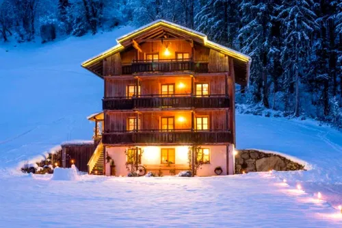 Ein charmantes Holzhaus mit mehreren Stockwerken, umgeben von schneebedeckten Bergen. Es strahlt in warmem Licht und hat dekorative Au&szlig;enlichter. Der Schnee liegt sanft auf dem Boden und es sind Lichtspuren im Vordergrund zu sehen.