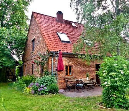 Ein charmantes Backsteinhaus mit rotem Ziegeldach. Vor der Terrasse stehen Tische und St&uuml;hle. Umgeben von &uuml;ppigem Gr&uuml;n und bunten Blumen.