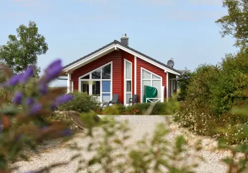 Rotes Haus mit modernen Fenstern und einer Veranda, umgeben von gr&uuml;nen Pflanzen und einem Kiesweg.