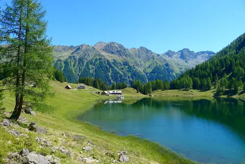 Eine idyllische Landschaft mit einem klaren See, umgeben von gr&uuml;nen Wiesen und hohen Bergen im Hintergrund. Fichten stehen am Ufer des Sees, w&auml;hrend vereinzelte H&uuml;tten auf der anderen Seite sichtbar sind. Der Himmel ist strahlend blau und sonnig.