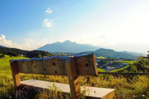 Eine Holzbank steht auf einem H&uuml;gel mit Blick auf Berge und ein Tal im Sonnenlicht. Die Landschaft ist gr&uuml;n und friedlich, mit Wolken im blauen Himmel.