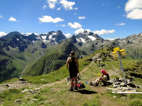Zwei Wanderer stehen auf einem Bergr&uuml;cken mit einer beeindruckenden Berglandschaft im Hintergrund. Eine Person ist mit einem Rucksack besch&auml;ftigt, w&auml;hrend die andere auf einem Felsen sitzt. Richtungsschilder zeigen verschiedene Wanderwege an. Die Szenerie ist klar und sonnig, mit schneebedeckten Gipfeln in der Ferne.