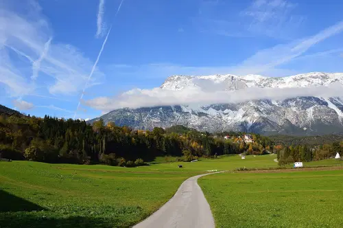 Eine malerische Landschaft mit gr&uuml;nen Wiesen, einem kurvigen Weg und schneebedeckten Bergen im Hintergrund. Der Himmel ist klar mit einigen Wolken und Kondensstreifen.