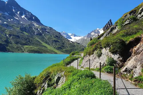 Ein malerischer Wanderweg entlang eines t&uuml;rkisfarbenen Sees, umgeben von gr&uuml;nen Wiesen und majest&auml;tischen Bergen mit schneebedeckten Gipfeln im Hintergrund. Der Himmel ist klar und blau.