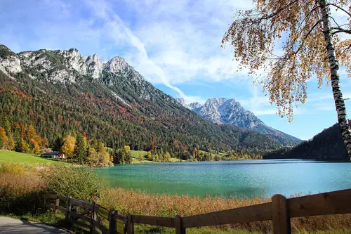 Ein malerischer Bergsee umgeben von hohen Bergen und buntem Laub. Ein Waldrand und eine Holzbank sind im Vordergrund sichtbar. Blaues Wasser reflektiert den klaren Himmel.