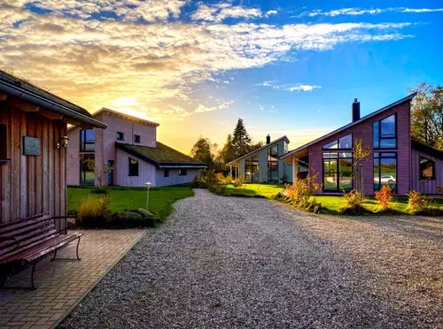 Helle, moderne Geb&auml;ude in l&auml;ndlicher Umgebung unter einem strahlend blauen Himmel mit vereinzelten Wolken. Die Sonne scheint und sorgt f&uuml;r eine warme Atmosph&auml;re. Ein Kiesweg f&uuml;hrt durch das Grundst&uuml;ck.