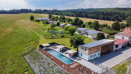 Luftaufnahme einer l&auml;ndlichen Unterkunft mit Schwimmbad, umgeben von gr&uuml;nen Wiesen und B&auml;umen. Mehrere Geb&auml;ude und G&auml;rten sind sichtbar, die eine einladende Umgebung schaffen.