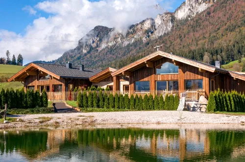 Zwei holzverkleidete H&auml;user stehen am Ufer eines ruhigen Sees, umgeben von gr&uuml;nen B&uuml;schen. Im Hintergrund erheben sich majest&auml;tische Berge unter einem blauen Himmel mit einigen Wolken.