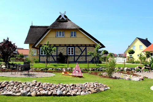 Ein gelbes, reetgedecktes Haus mit dekorativen Elementen, umgeben von einem gepflegten Garten mit Steinen und Sitzgelegenheiten. Im Hintergrund sind weitere Wohnh&auml;user und ein klarer Himmel zu sehen.