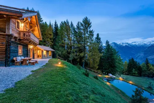 Eine rustikale Holzh&uuml;tte steht in einer malerischen Landschaft, umgeben von B&auml;umen und einem kleinen Teich. Das Licht der D&auml;mmerung wirft einen warmen Schein auf die Struktur, w&auml;hrend die bergige Kulisse im Hintergrund sichtbar ist.