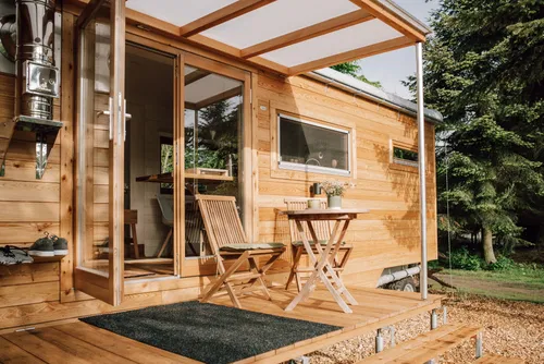 Ein minimalistisches Holzhaus mit offener Terrasse, ausgestattet mit einem kleinen Tisch und zwei St&uuml;hlen. Der Zugang erfolgt durch gro&szlig;e Glast&uuml;ren, umgeben von Natur.