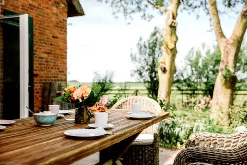 Ein gedeckter Tisch auf einer Terrasse, umgeben von B&auml;umen und einer gr&uuml;nen Wiese. Eine Blumenvase mit bunten Blumen steht in der Mitte, daneben liegen Teller und eine Sch&uuml;ssel mit Snacks.