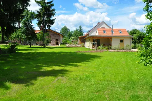 Zwei H&auml;user mit roten D&auml;chern stehen in einem gr&uuml;nen Garten. Umgeben von B&auml;umen und einem blauen Himmel mit einigen Wolken.