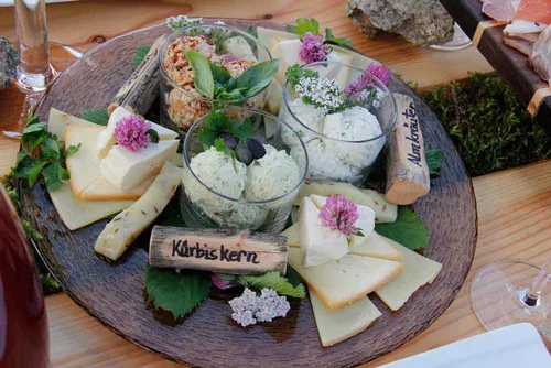 Ein rustikeller Teller mit verschiedenen K&auml;sesorten, Aufstrichen und Kr&auml;utern. Die K&auml;sest&uuml;cke sind liebevoll angerichtet und einige mit essbaren Blumen verziert. Holzkarten beschriften die verschiedenen Sorten.