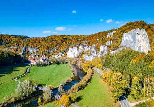 Eine malerische Landschaft mit bunten Herbstb&auml;umen, einem ruhigen Fluss und steilen Felsen. Im Hintergrund sind ein paar Geb&auml;ude zu sehen, umgeben von viel Gr&uuml;n und farbenfrohen Bl&auml;ttern. Der Himmel ist klar und blau.