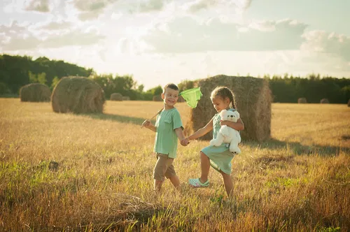 Zwei Kinder halten H&auml;ndchen und laufen fr&ouml;hlich durch ein Getreidefeld. Eines der Kinder tr&auml;gt ein Netz und das andere h&auml;lt ein Pl&uuml;schtier. Im Hintergrund sind Heuballen und ein klarer Himmel zu sehen.