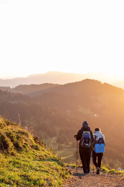Zwei Wanderer mit Rucks&auml;cken gehen einen Pfad entlang, w&auml;hrend die Sonne hinter den Bergen aufgeht. Die Landschaft ist gr&uuml;n und bewaldet, mit sanften H&uuml;geln im Hintergrund.