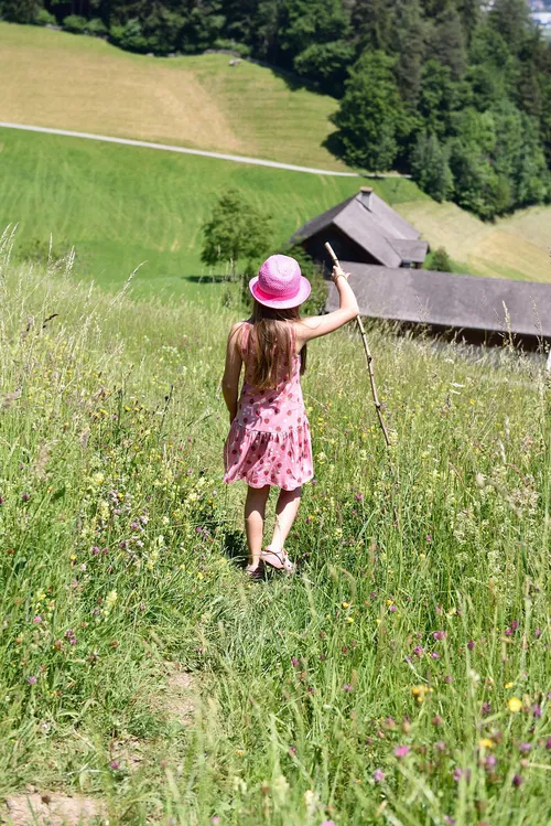 Ein M&auml;dchen mit einem rosa Hut und einem blumigen Kleid steht auf einem bl&uuml;henden Feld und h&auml;lt einen Stock. Im Hintergrund sind eine Scheune und bewaldete H&uuml;gel sichtbar.