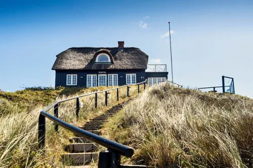Ein traditionelles Reetdachhaus steht auf einer Anh&ouml;he, umgeben von wuscheligen Grasfl&auml;chen. Eine steinerne Treppe f&uuml;hrt zu dem Haus, das eine gro&szlig;e Fensterfront hat. Der Himmel ist blau mit einigen Wolken.