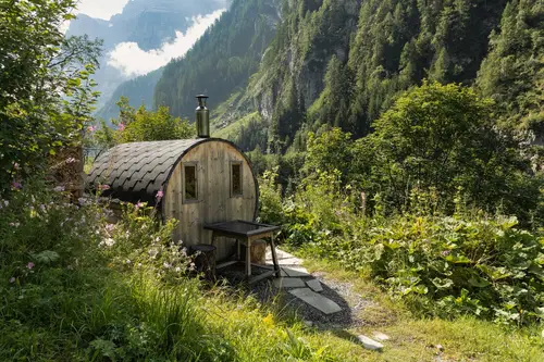 Eine kleine h&ouml;lzerne H&uuml;tte steht malerisch in einer gr&uuml;nen Landschaft, umgeben von B&auml;umen und Bergen. Der Weg zur H&uuml;tte ist steinig, und bunte Blumen bl&uuml;hen im Vordergrund.