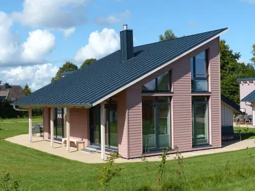 Ein modernes, eink&ouml;mmliches Haus mit schr&auml;g verlaufendem Dach, gro&szlig;en Fenstern und einer Holzfassade in Rosat&ouml;nen. Das Grundst&uuml;ck ist gr&uuml;n und von einer Wiese umgeben. Helle Wolken zieren den blauen Himmel.