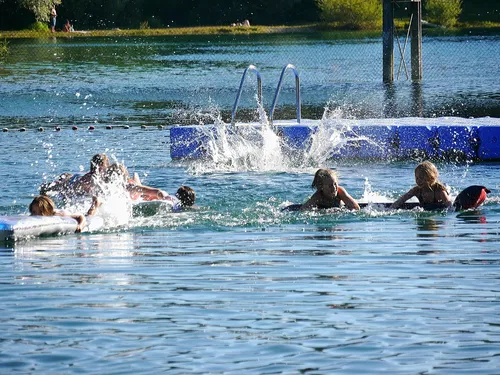 Kinder und Erwachsene schwimmen und planschen in einem klaren Wasser an einem warmen Tag. In der Mitte des Sees sind Spritzer zu sehen, w&auml;hrend einige Personen auf einem Schwimmbrett liegen. Im Hintergrund ist eine Badeleiter und einige B&auml;ume sichtbar.