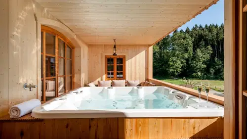 Entspannender Whirlpool auf einer Holzterrasse, umgeben von Fenstern. Im Hintergrund gr&uuml;ne B&auml;ume und eine ruhige Landschaft. Zwei Gl&auml;ser Sekt stehen bereit.