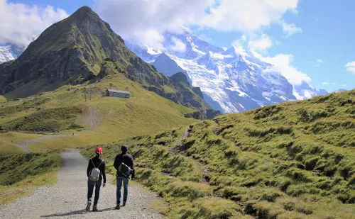 Zwei wandernde Personen auf einem Pfad in den Bergen, umgeben von gr&uuml;nem Gras und schneebedeckten Gipfeln, unter einem klaren blauen Himmel.